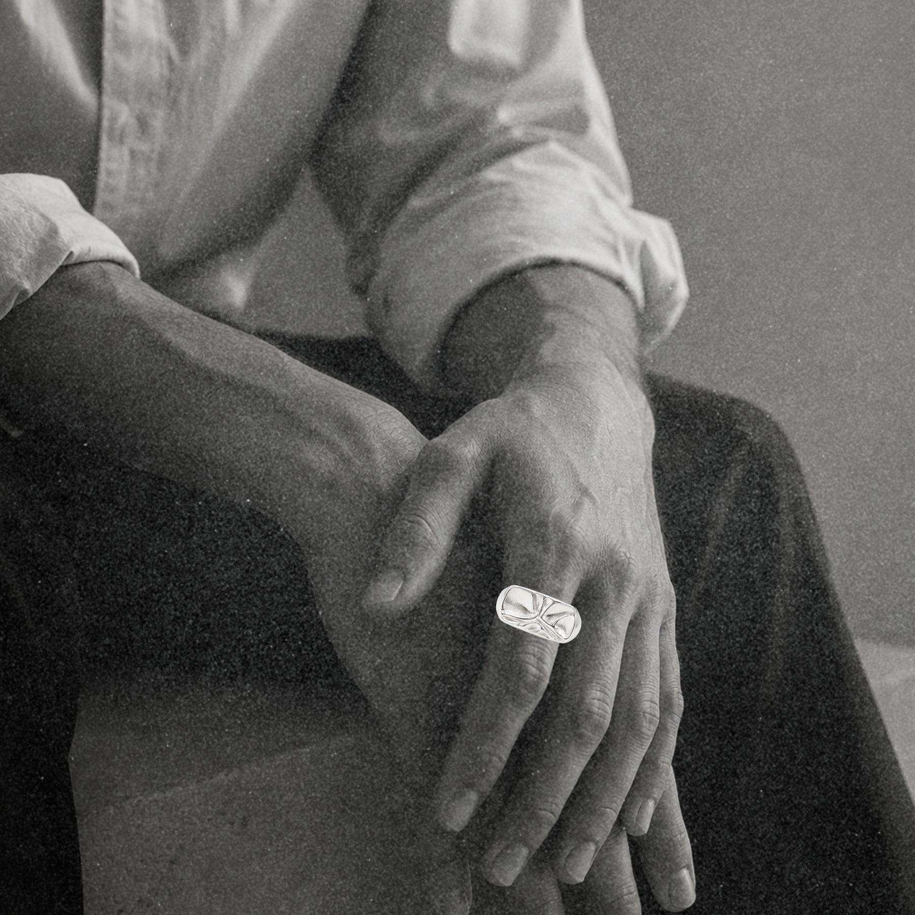 A close-up black and white photograph of a person's hands resting on their lap, wearing a silver signet ring with an abstract rippled pattern on the face.