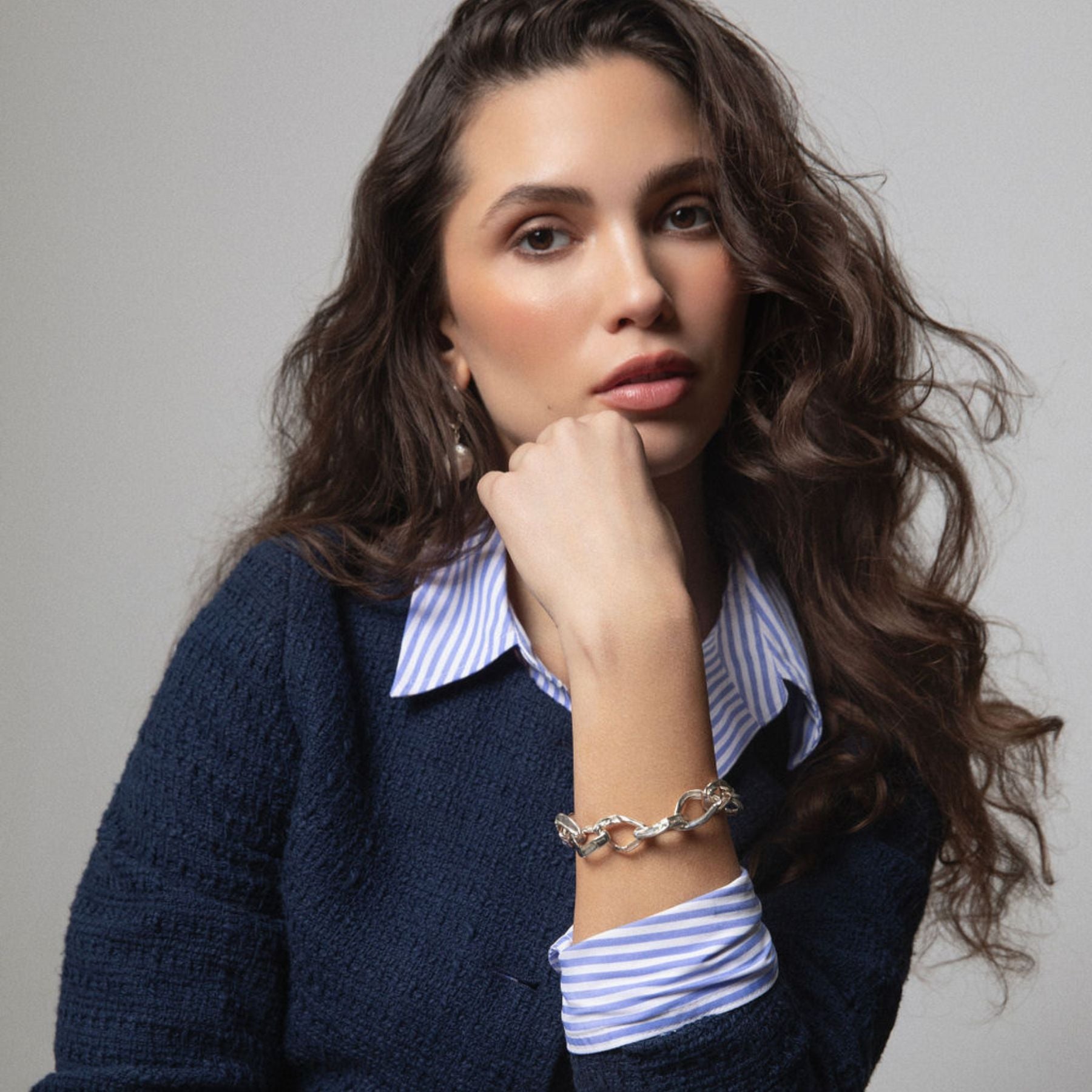 A woman with long wavy brown hair poses in a navy blue knit cardigan over a blue and white striped collared shirt, wearing a silver chain link bracelet by Jessica Liu on her wrist against a neutral gray background.
