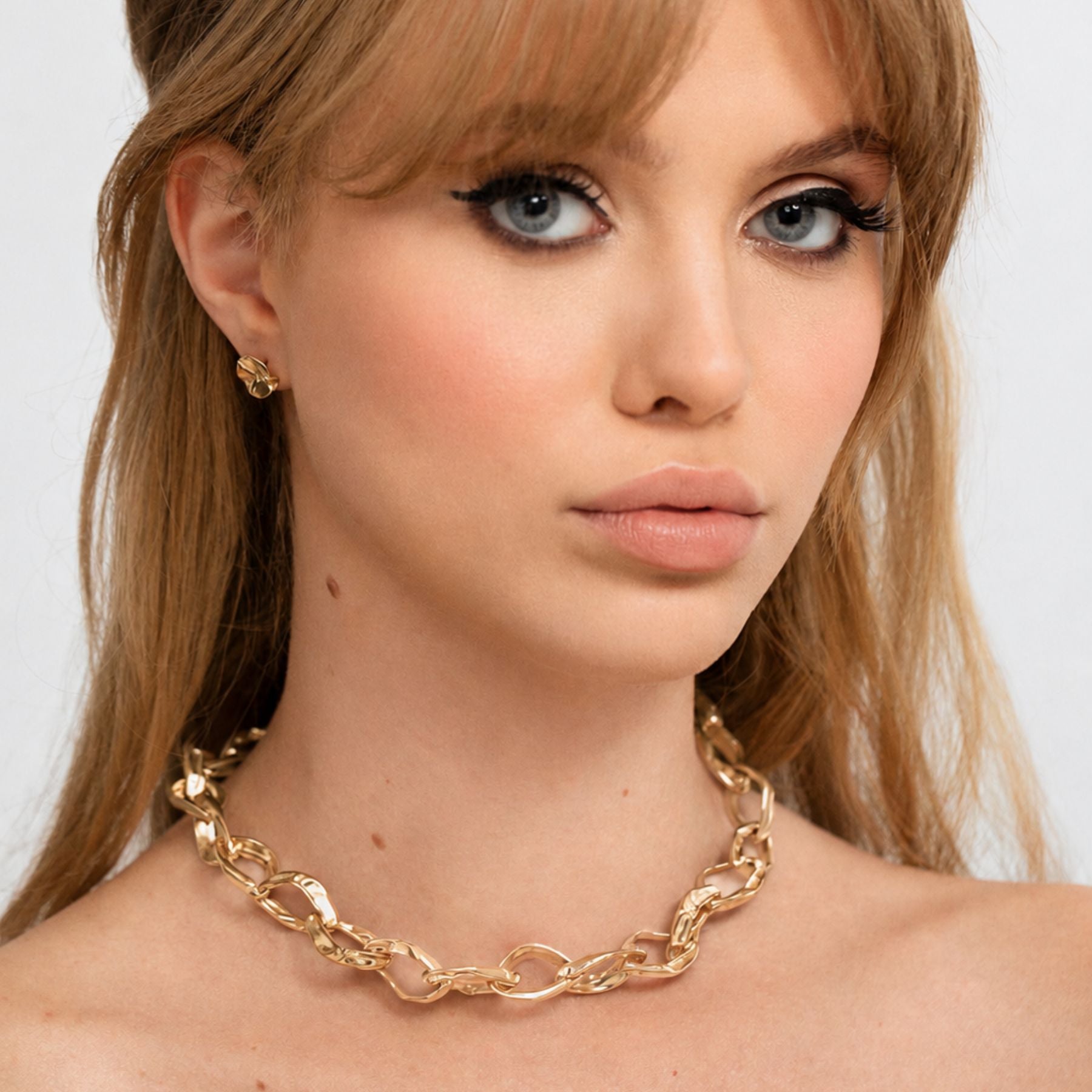 A close-up of a woman wearing a chunky gold chain necklace and a matching small gold stud earring by Jessica Liu against a light gray background.