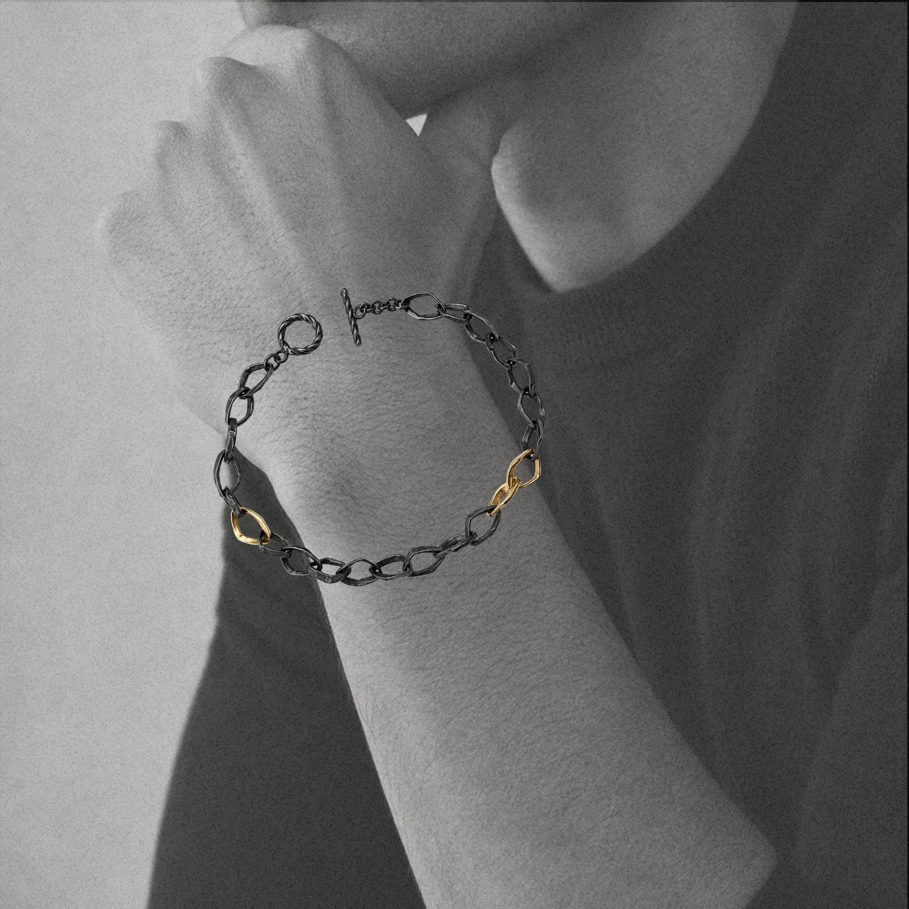 A delicate chain bracelet by Jessica Liu with a black oxidized finish and two small gold infinity links, shown worn on a person's wrist in a close-up grayscale lifestyle shot.