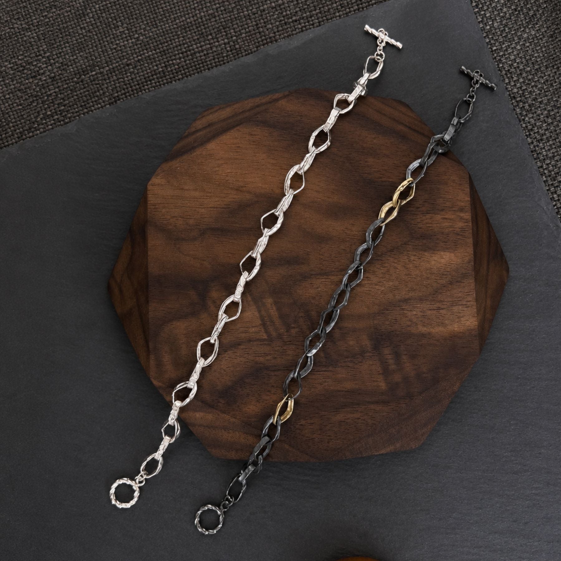 Two chain bracelets by Jessica Liu displayed on a dark wooden block over a dark gray slate surface. One bracelet is silver with textured oval links and a toggle clasp, and the other is black with small 18k gold accent links and matching toggle clasp.