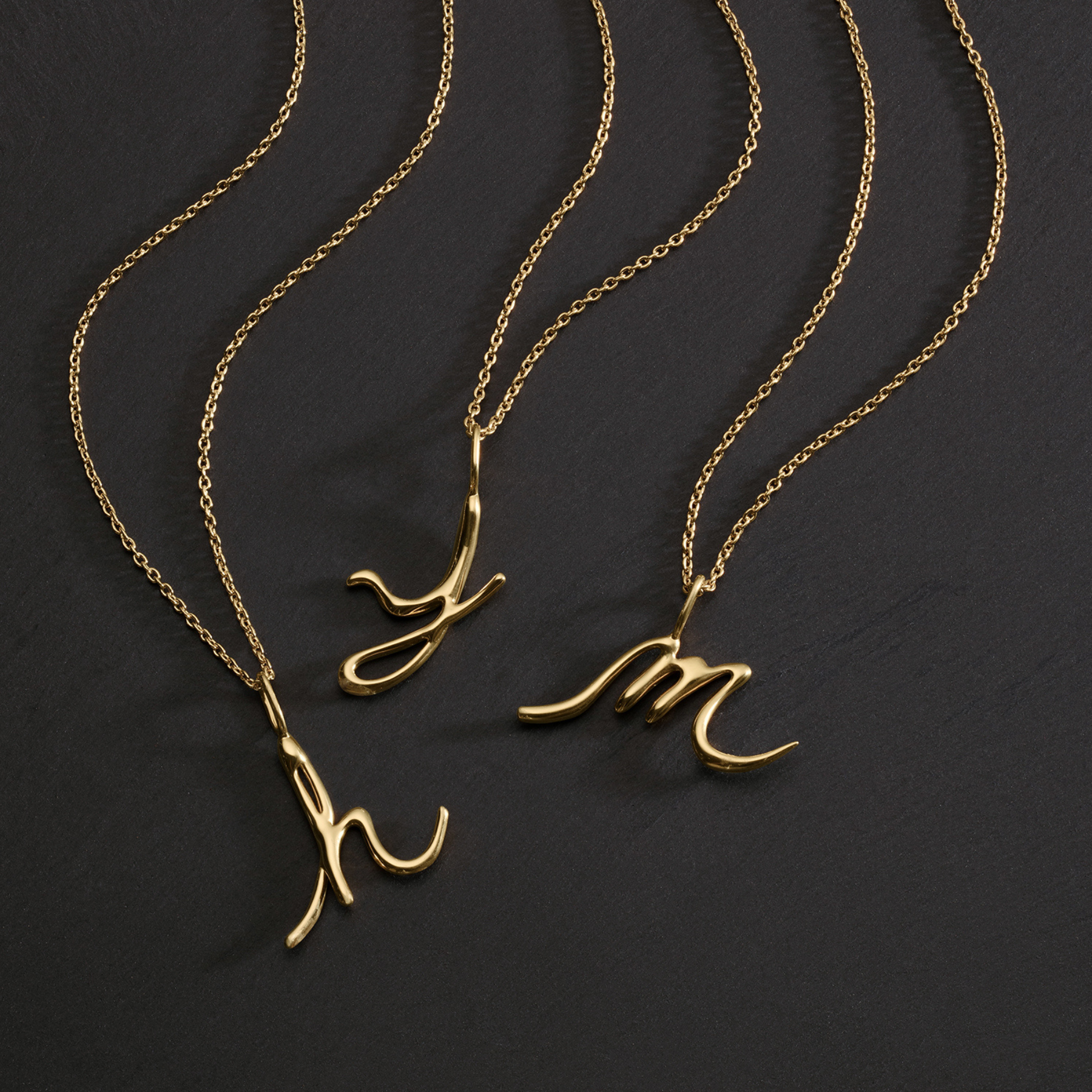 14k gold alphabet initial necklaces in letters h, j, and m against a black background by jessica liu