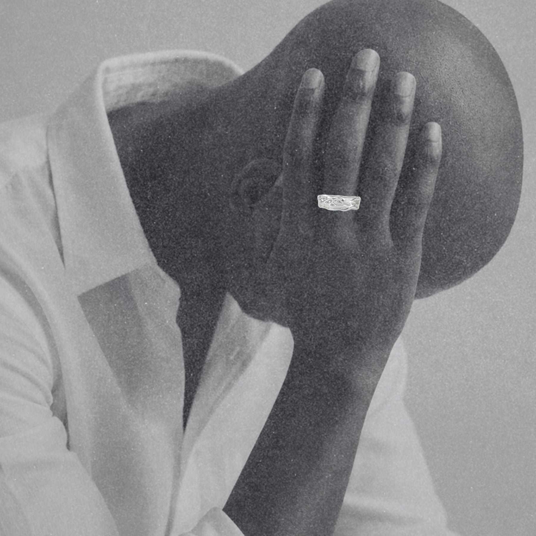 A black and white close-up of a person in a white collared shirt holding their head in their hands, with a textured silver ring by Jessica Liu on one finger.
