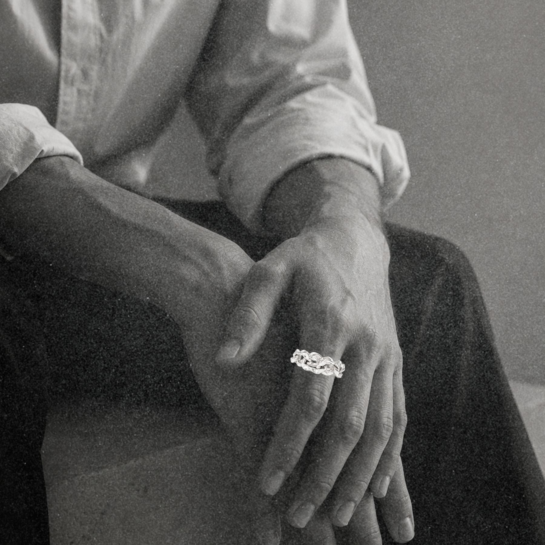 A close-up black and white photo of a person seated with hands crossed, wearing a silver chain ring by Jessica Liu on one finger. 