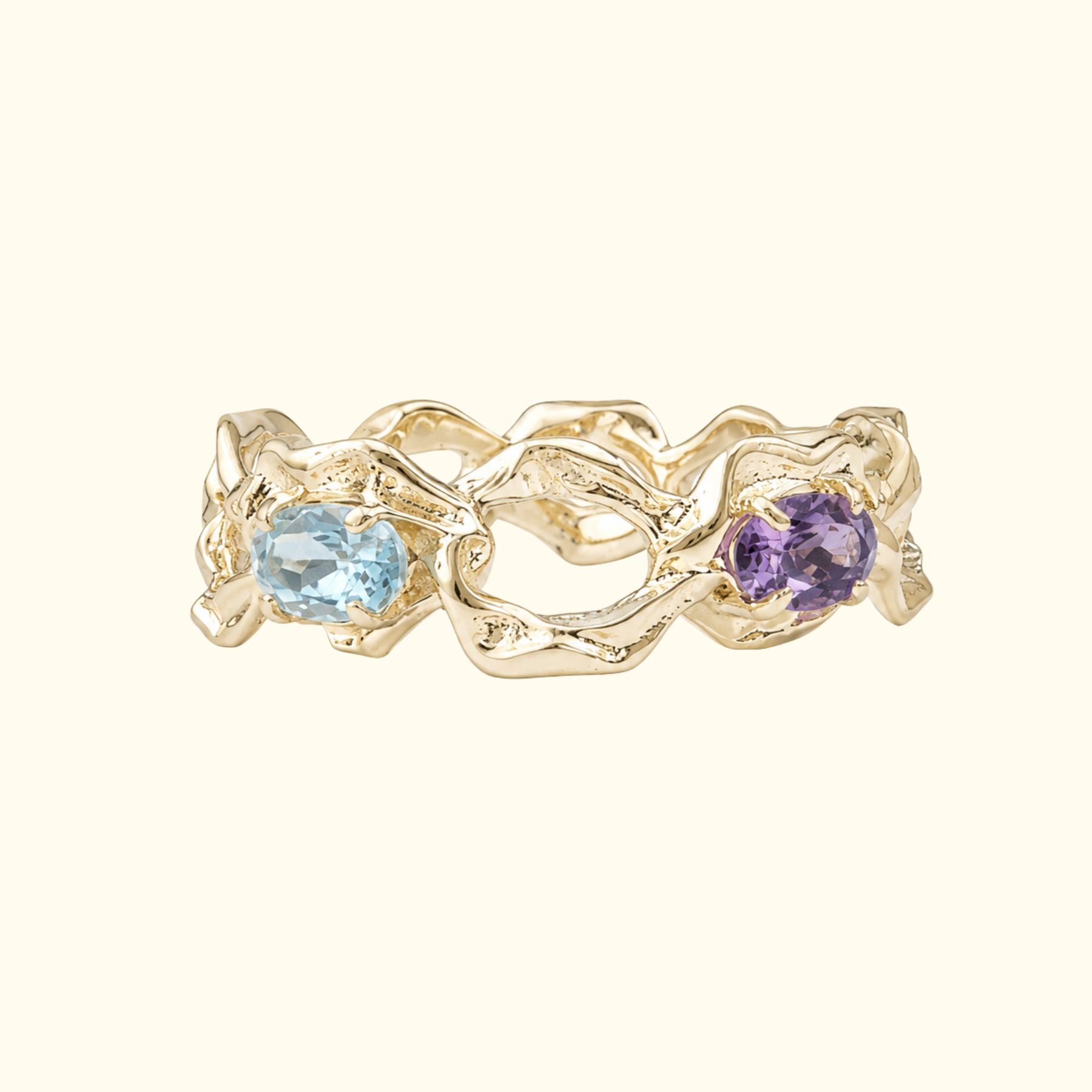 A gold ring by Jessica Liu with an organic, wavy textured band showing two oval gemstones, one light blue and one purple, set against a solid cream background.