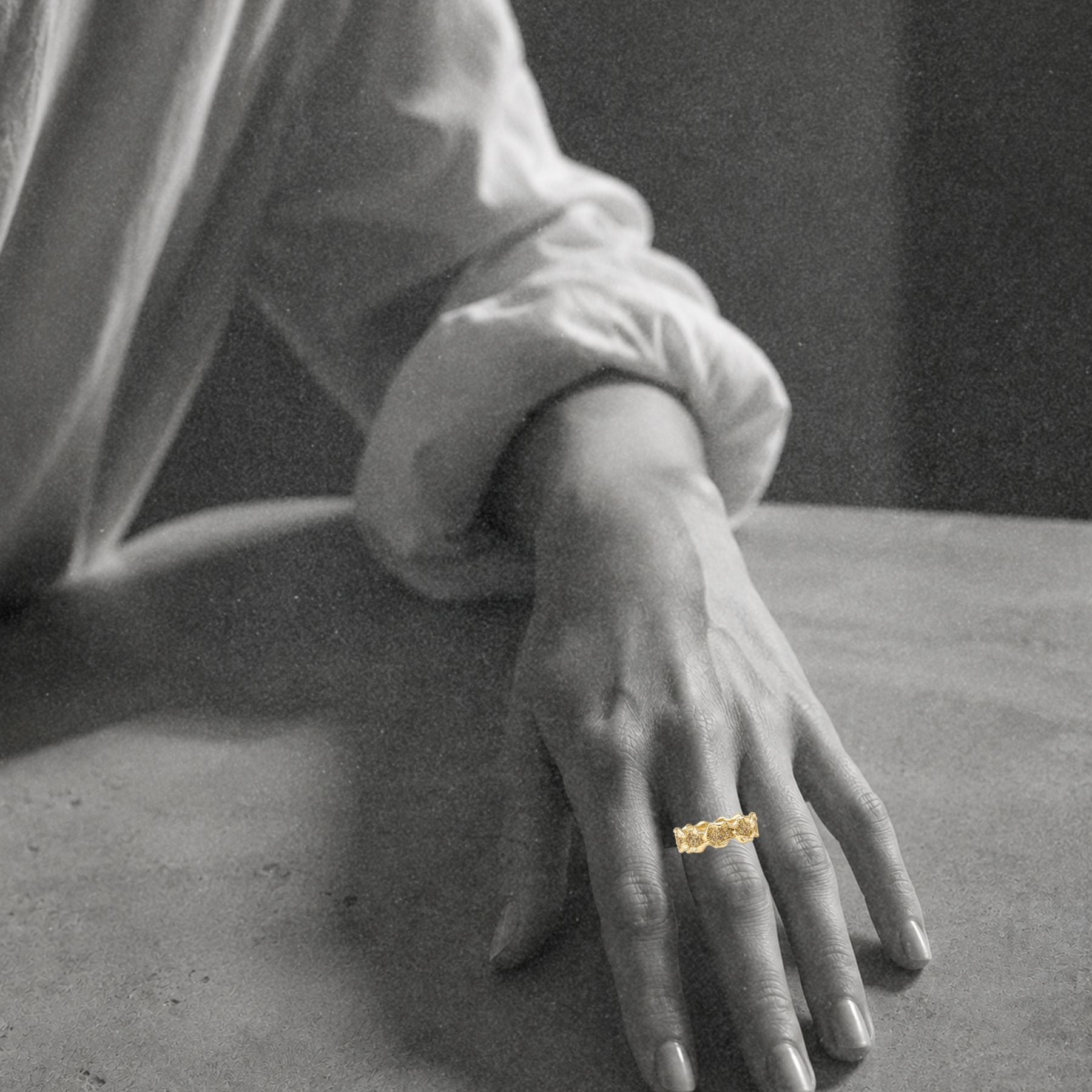 A close-up of a person's hand resting on a surface, wearing a textured gold ring with champagne diamonds by Jessica Liu on one finger. The image is in black and white, emphasizing the skin tones and the gold ring, with a blurred background.