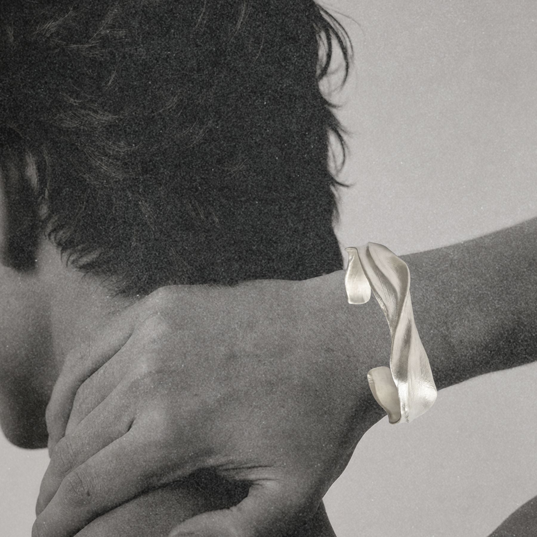 A close-up black and white photo of a person's hand and neck, wearing a silver twisted ribbon-style cuff by Jessica Liu on the wrist.