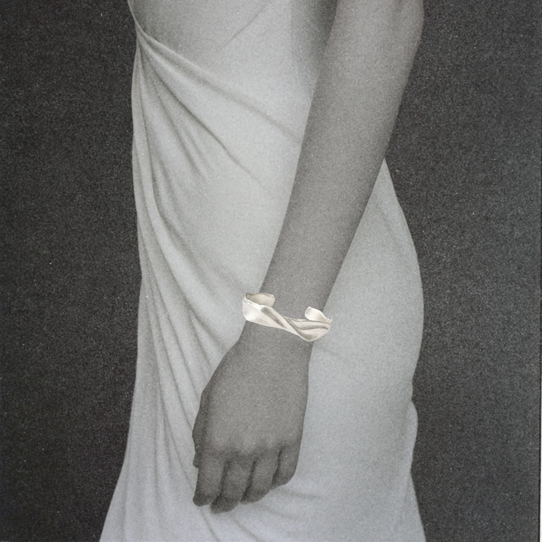 A close-up of a person wearing a rippled cuff silver bracelet by Jessica Liu on their wrist, dressed in a white draped dress against a dark textured background.